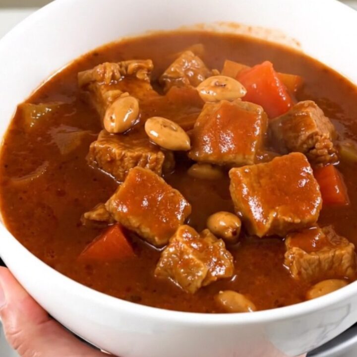 A close-up of a steaming bowl of pork peanut stew with vibrant carrots and a rich, glossy sauce.