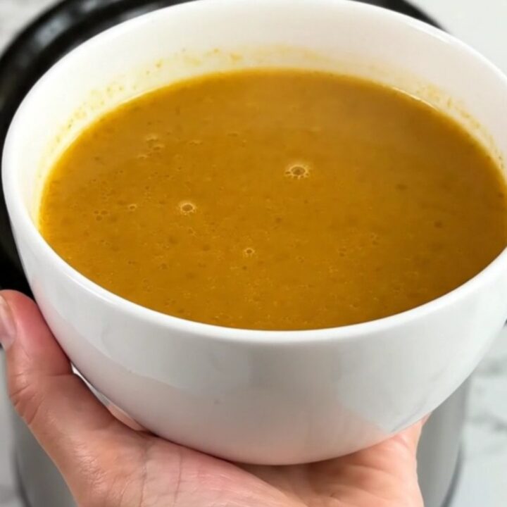 A white ceramic bowl filled with velvety, golden pumpkin and red lentil soup, steaming gently.