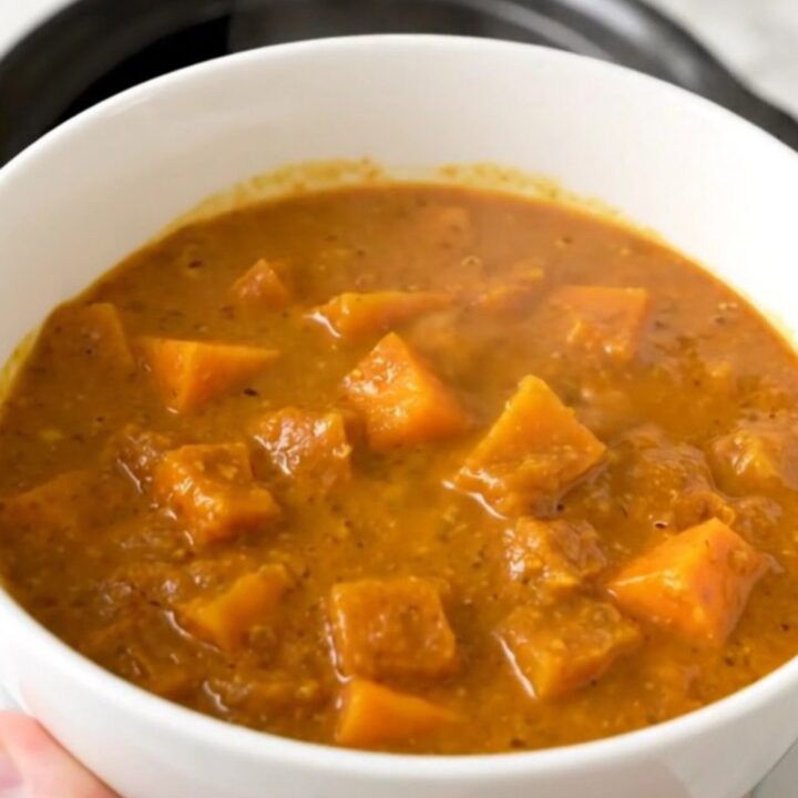 A top-down view of a black slow cooker filled with raw red lentils, diced butternut squash, and coconut milk.
