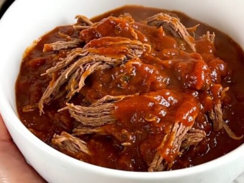 A close-up of a white bowl filled with glossy, tender shredded beef chili with a rich red tomato sauce.