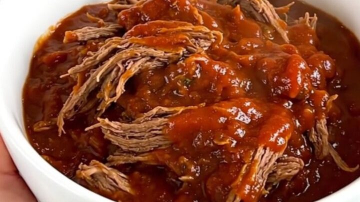A close-up of a white bowl filled with glossy, tender shredded beef chili with a rich red tomato sauce.