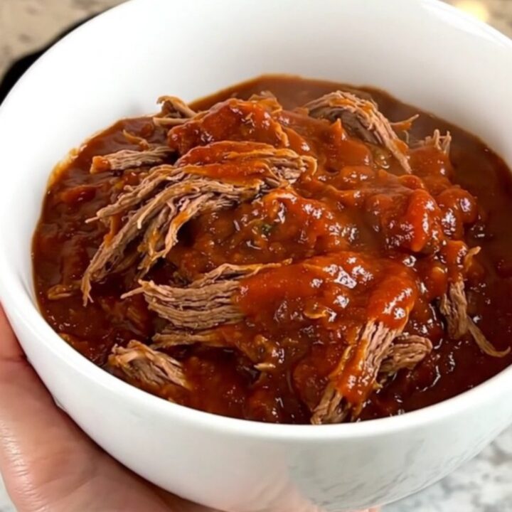 A close-up of a white bowl filled with glossy, tender shredded beef chili with a rich red tomato sauce.
