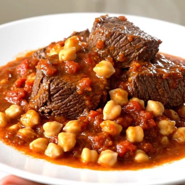 A top-down view of raw beef chuck pieces, chickpeas, and crushed tomatoes in a black slow cooker on a speckled countertop.