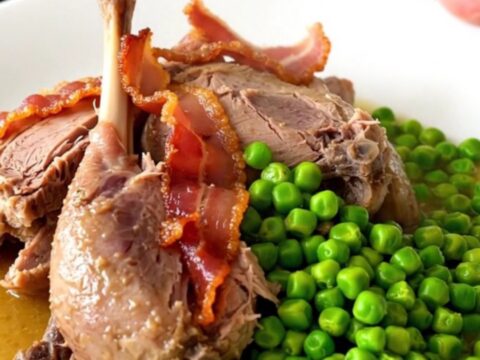 A rustic slow-cooked guinea fowl dish with bacon lardons and bright green peas in a dark oval slow cooker.