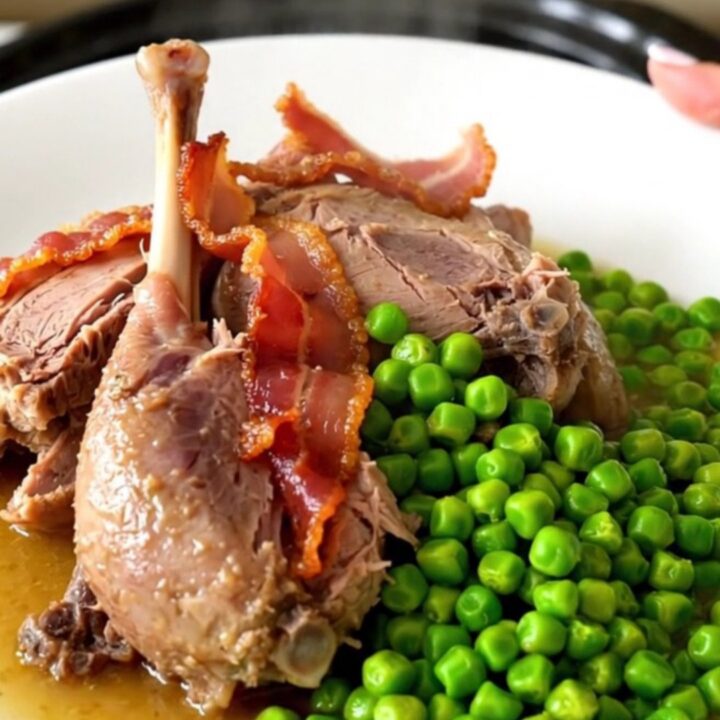 A rustic slow-cooked guinea fowl dish with bacon lardons and bright green peas in a dark oval slow cooker.