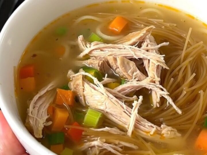 Bol de soupe de poulet fumante avec vermicelles et légumes colorés dans une cuisine lumineuse.