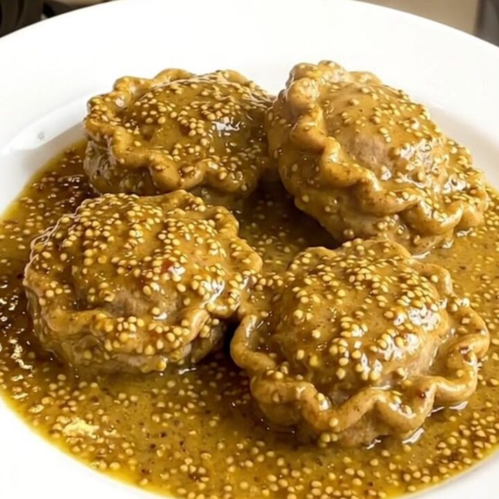 A close-up of tender veal paupiettes coated in a creamy, glossy mustard sauce on a white plate.