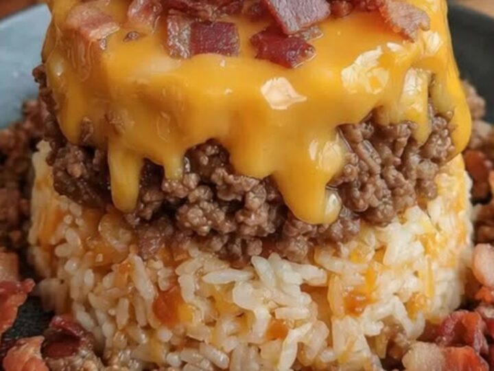 A bacon cheeseburger rice stack on a dark plate, featuring layers of rice, beef, melted cheddar, and bacon.