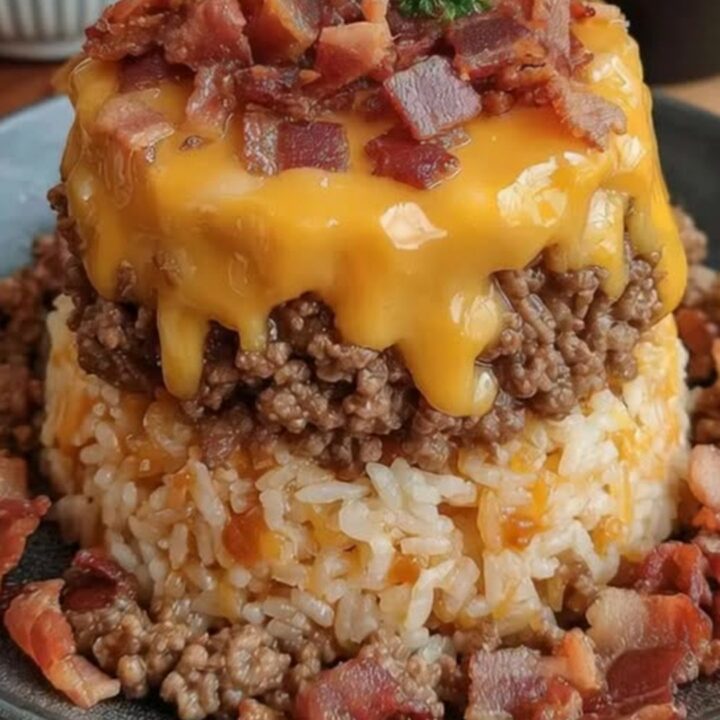 A bacon cheeseburger rice stack on a dark plate, featuring layers of rice, beef, melted cheddar, and bacon.