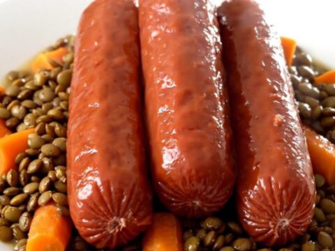 A steaming plate of slow-cooked lentils with diced carrots and sliced frankfurters on a white plate.