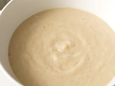 A steaming, glossy white bowl of turnip and leek cream soup set against a soft-blurred cream kitchen background.
