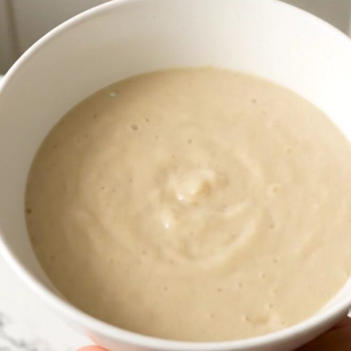 A steaming, glossy white bowl of turnip and leek cream soup set against a soft-blurred cream kitchen background.