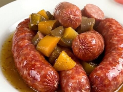 A white plate filled with cooked cervelas sausages, sliced potatoes, leeks, and carrots in a mustard broth.