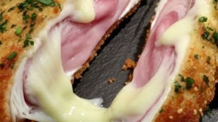 A golden, crispy Chicken Cordon Bleu cut in half to show melted Swiss cheese and ham on a dark slate background.