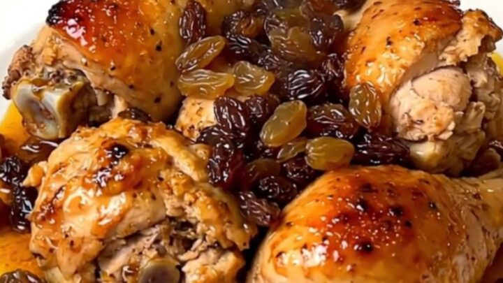 A close-up view of tender chicken thighs with golden raisins in a savory sauce on a white plate.