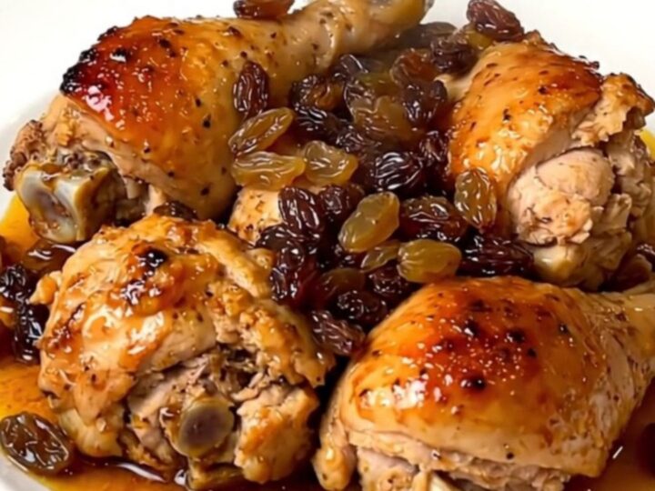 A close-up view of tender chicken thighs with golden raisins in a savory sauce on a white plate.
