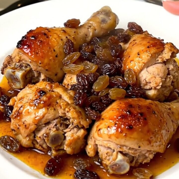 A close-up view of tender chicken thighs with golden raisins in a savory sauce on a white plate.
