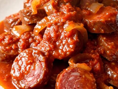 A top-down view of a slow cooker filled with slices of andouille sausage, onions, and tomato sauce ready for cooking.
