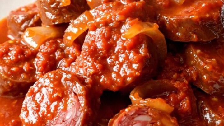 A top-down view of a slow cooker filled with slices of andouille sausage, onions, and tomato sauce ready for cooking.