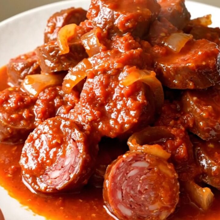 A top-down view of a slow cooker filled with slices of andouille sausage, onions, and tomato sauce ready for cooking.