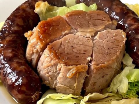 A top-down view of a slow cooker filled with pork shoulder, sliced sausages, and shredded cabbage.