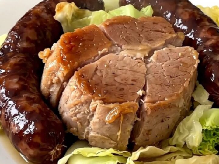 A top-down view of a slow cooker filled with pork shoulder, sliced sausages, and shredded cabbage.