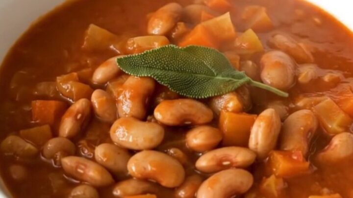 A rustic bowl of steaming borlotti bean stew topped with fresh sage leaves in a bright kitchen.