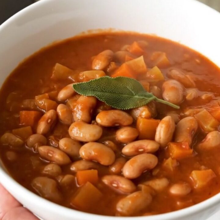 A rustic bowl of steaming borlotti bean stew topped with fresh sage leaves in a bright kitchen.
