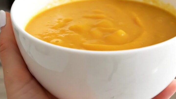 A white bowl filled with smooth, vibrant orange pumpkin and leek soup against a blurred kitchen background.