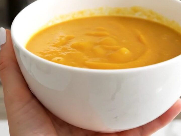 A white bowl filled with smooth, vibrant orange pumpkin and leek soup against a blurred kitchen background.