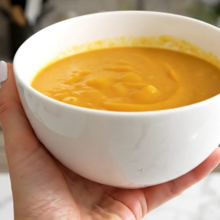 A white bowl filled with smooth, vibrant orange pumpkin and leek soup against a blurred kitchen background.