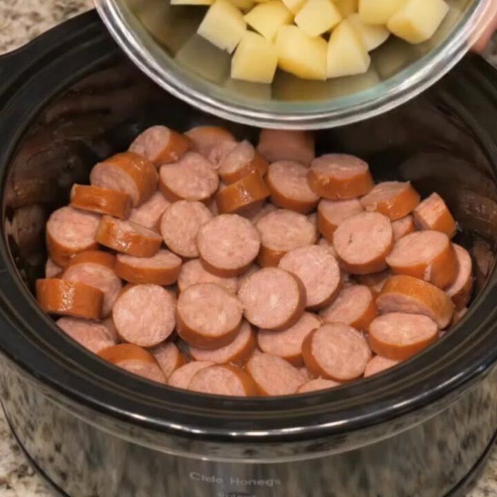 A fork scooping a tender potato cube and a glossy browned sausage slice from a steaming slow cooker.