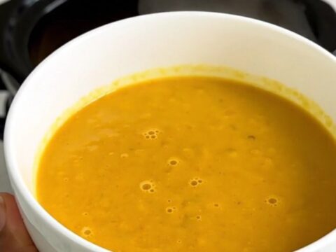 A bowl of vibrant orange red kuri squash soup topped with shaved Comté cheese and crispy bacon bits.