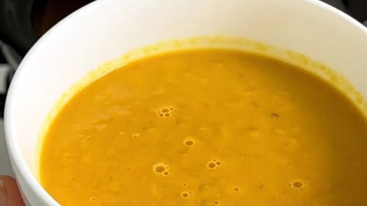 A bowl of vibrant orange red kuri squash soup topped with shaved Comté cheese and crispy bacon bits.