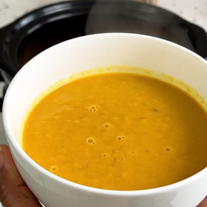 A bowl of vibrant orange red kuri squash soup topped with shaved Comté cheese and crispy bacon bits.