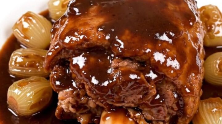A tender, glazed pork shank served on a white plate with pearl onions and carrots in a rich red wine sauce.