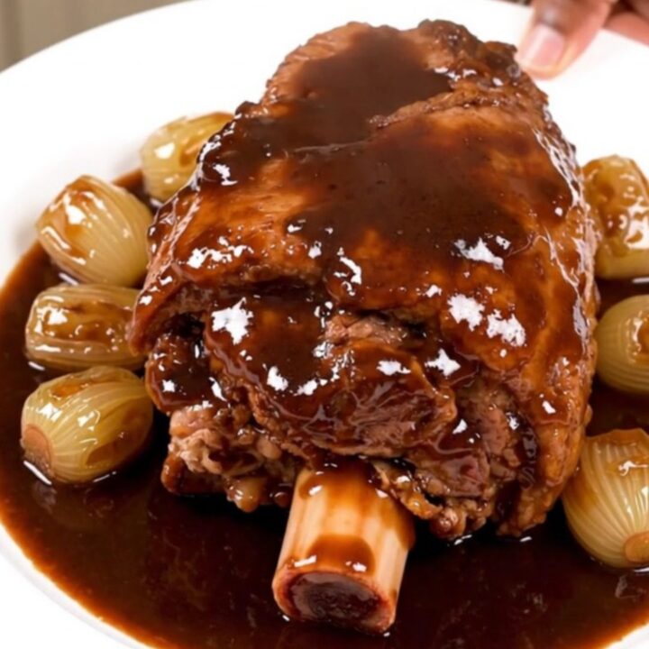 A tender, glazed pork shank served on a white plate with pearl onions and carrots in a rich red wine sauce.