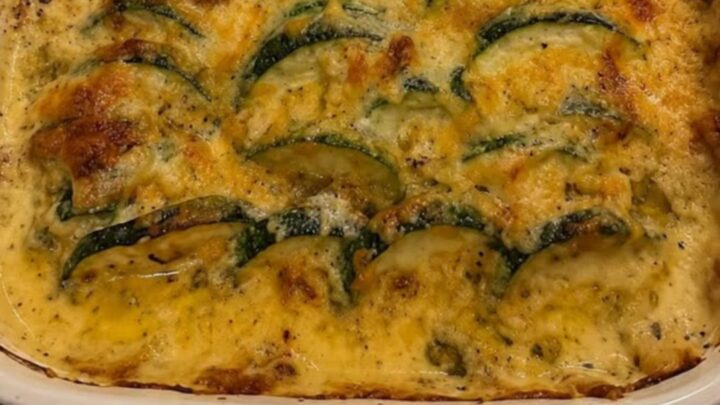 A steaming cheesy zucchini and ground beef casserole in a white baking dish with red handles.