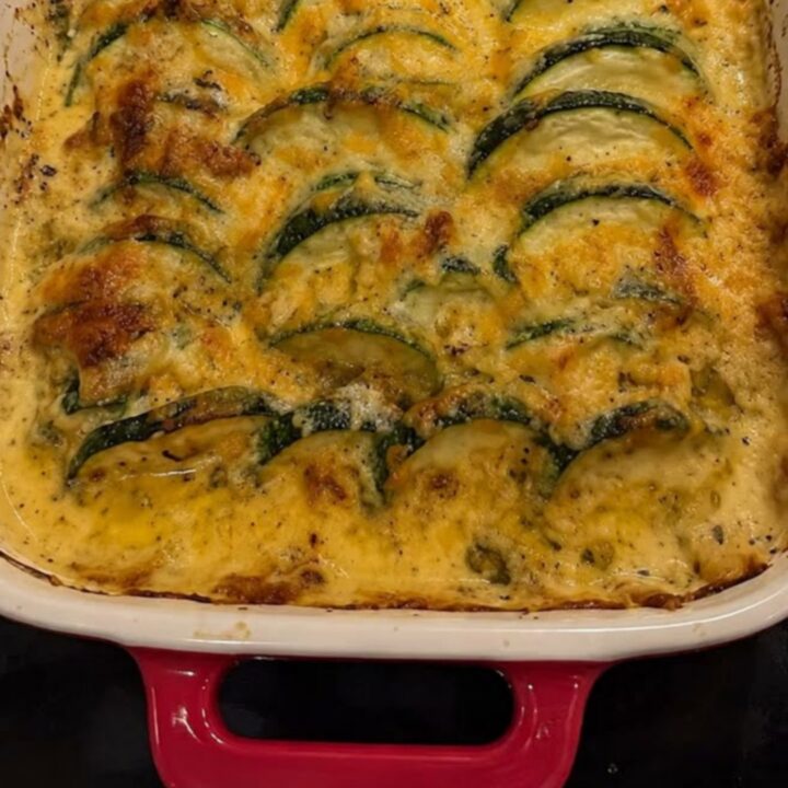 A steaming cheesy zucchini and ground beef casserole in a white baking dish with red handles.