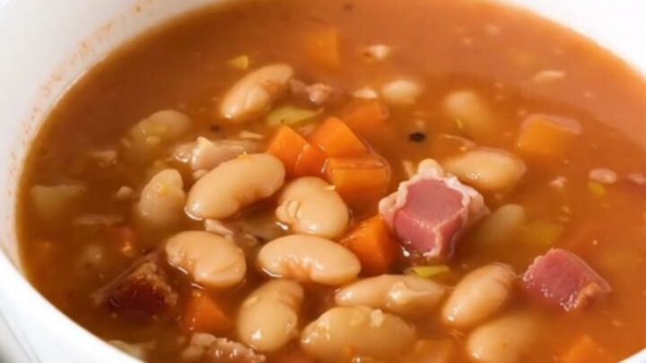 A steaming bowl of mountain minestrone soup with bacon and vegetables in a white ceramic bowl.