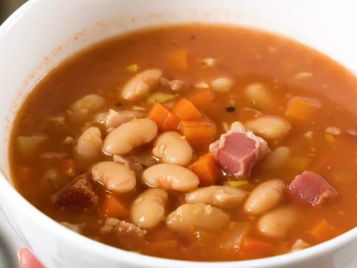 A steaming bowl of mountain minestrone soup with bacon and vegetables in a white ceramic bowl.