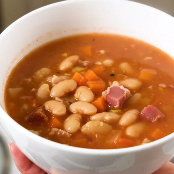 A steaming bowl of mountain minestrone soup with bacon and vegetables in a white ceramic bowl.