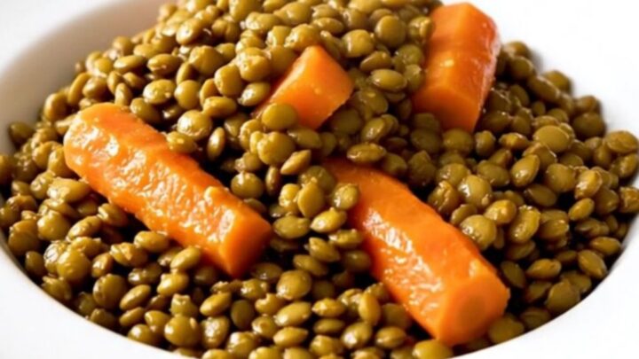 A close-up of a bowl filled with slow-cooked green lentils, carrots, and onions in a savory, glossy mustard broth.