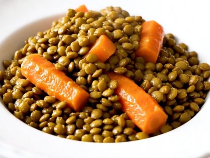 A close-up of a bowl filled with slow-cooked green lentils, carrots, and onions in a savory, glossy mustard broth.