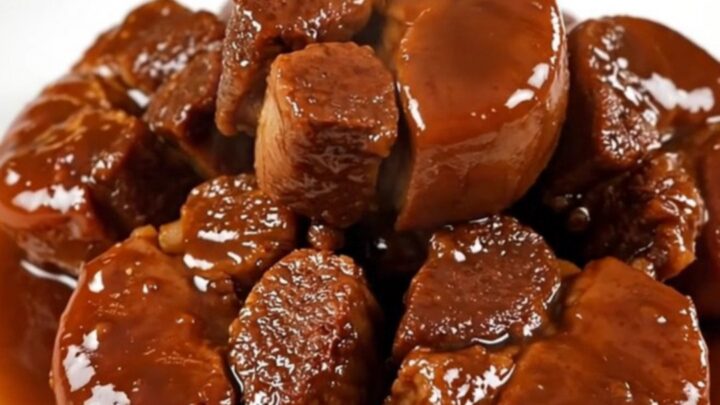A close-up of glossy, fork-tender braised pork cheeks served on a clean white round plate with sliced carrots.