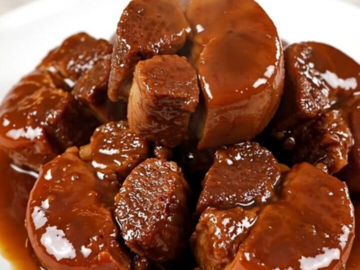 A close-up of glossy, fork-tender braised pork cheeks served on a clean white round plate with sliced carrots.