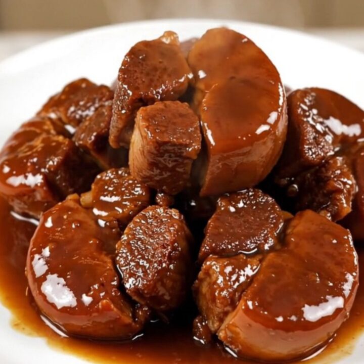 A close-up of glossy, fork-tender braised pork cheeks served on a clean white round plate with sliced carrots.