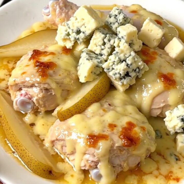 A close-up of tender chicken thighs coated in a rich, creamy Fourme d'Ambert blue cheese sauce with pear wedges.