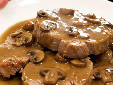 A tender, slow-cooked pork roast topped with a creamy mushroom and shallot sauce on a white plate.