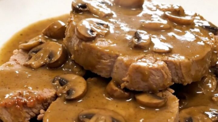 A tender, slow-cooked pork roast topped with a creamy mushroom and shallot sauce on a white plate.